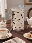 Ivory Blossoms jug cover in cream fitted on a glass water pitcher styled on a brown table with tea china and macarons.
