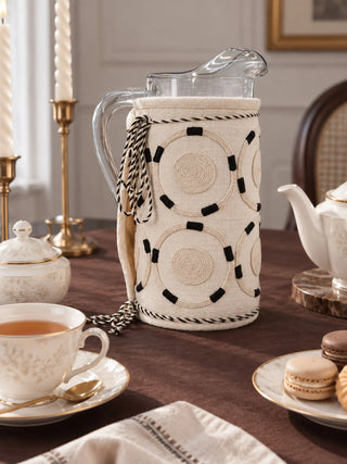 Ivory Blossoms jug cover in cream fitted on a glass water pitcher styled on a brown table with tea china and macarons.