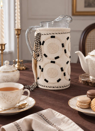 The Ivory Blossoms jug cover in Ivory, view-diningtable on a glass pitcher beside a teapot and macarons.