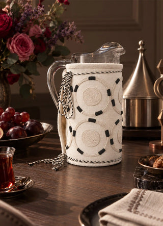 The Ivory Blossoms jug cover in Ivory, view-eveningtable on a glass pitcher beside roses and fruit.