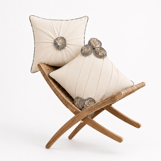 The Ivory Gardenia Draped Floral cushion set styled on a woven rattan bench, showing a round pintuck-pleated cushion with a central gardenia-style rosette and a diagonally pleated cushion with clustered rosettes, both in ivory cotton-linen.