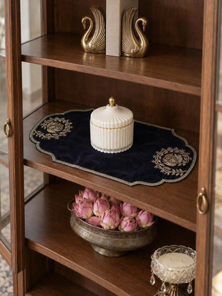 Jashn navy table mat with gold embroidery under a white ribbed lidded jar on a dark wood cabinet shelf, with brass swan bookends above and a bowl of pink lotus buds below.