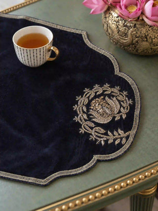 A close-up of the Jashn table mat’s gold wreath embroidery with a sequin-centred flower, and its scalloped metallic braid edge, beside a cup of tea.
