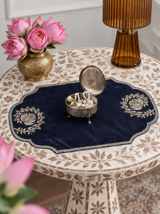 Jashn navy table mat on a round inlaid side table under a silver trinket box of pearls, with a brass vase of pink lotus flowers and an amber glass lamp.
