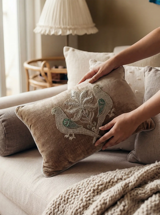 A lifestyle view of the hand-embroidered jewelled parrot cushion being styled on a neutral sofa.