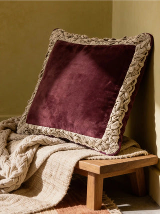 A Jhalak ivory and maroon cushion resting on a rustic wooden bench with a warm neutral throw.