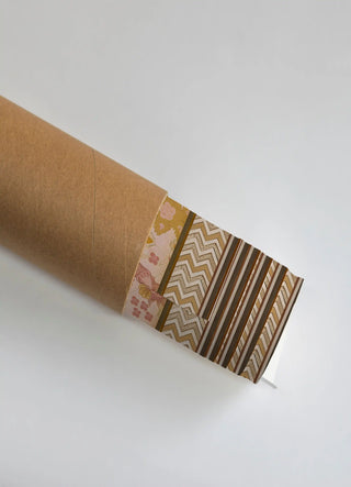 The Kahani Indian Art Theme wall art print partially unrolled from its kraft paper shipping tube, showing a rounded blossom floral panel beside a chevron zigzag border and diagonal stripe banding in pink, mustard and gold.