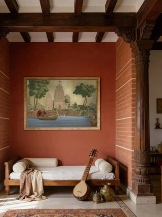 Kashi Reverie temple and swan boat wall art in antique sepia, framed in pale gold on a terracotta wall above a wooden daybed with bolsters and a veena in a beamed living room.