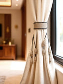 A luxury cream linen curtain held by a Kaya handcrafted jute tieback featuring a woven band and multiple small tassels.