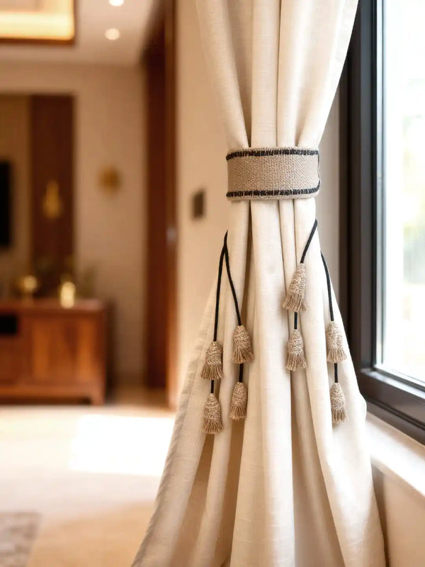 A luxury cream linen curtain held by a Kaya handcrafted jute tieback featuring a woven band and multiple small tassels.