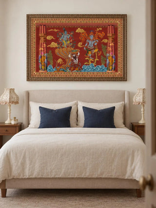 Keshava landscape wall art in an ornate bronze-toned frame above an upholstered bed with cream linen, two navy cushions and table lamps with floral shades.