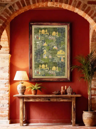 Krishna Leela pavilion and garden wall art in forest green and gold, framed in weathered wood on a deep red wall above a rustic console with a ceramic lamp and potted palm in a brick-arched entryway.