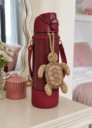 The Kurma charm in Gold, view-waterbottle hanging from a burgundy water bottle strap.