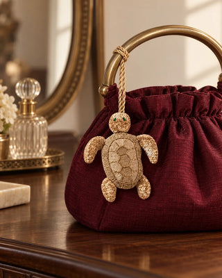 Kurma gold embroidered turtle charm tied to a burgundy silk potli bag with an antique brass handle.