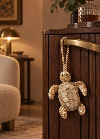 A hand-beaded Kurma turtle charm in gold and cream, its shell worked in sequins and beads, hung by rope from a brass cabinet handle