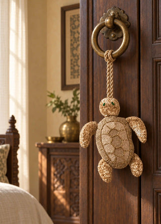 A hand-beaded Kurma turtle charm in gold and cream, its shell worked in sequins and beads, tied by rope to a brass ring door knocker.