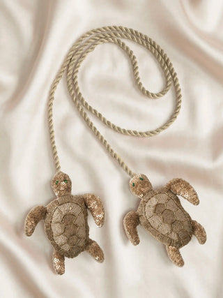 A flat-lay view of the Kurma turtle tieback styled on a shimmering champagne silk fabric.