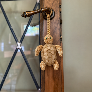 The Kurma Turtle Gift Charm in Champagne Gold, hung from a wooden door handle by its braided cord, showing the sequinned shell and green bead eyes.