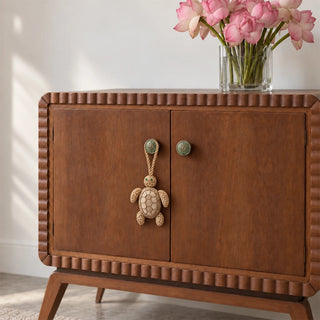The Kurma Turtle Gift Charm hanging from a green knob on a walnut-toned cabinet beside a glass vase of pink blooms.