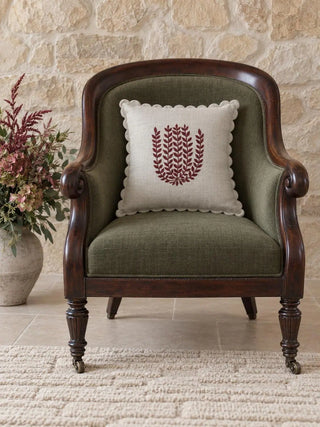 Leafora cushion cover with burgundy leaf-sprig embroidery and a scalloped edge on an olive-green upholstered armchair with a carved dark wood frame, against a pale stone wall.