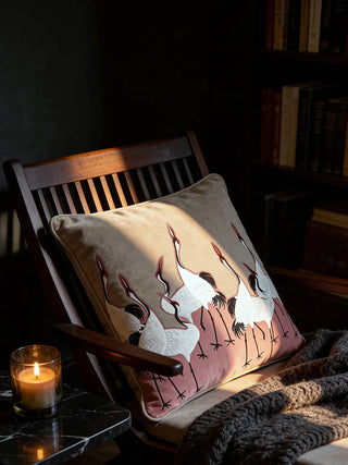 The Sedge Pink crane cushion styled on a dark wooden armchair in a moody, sunlit library setting.