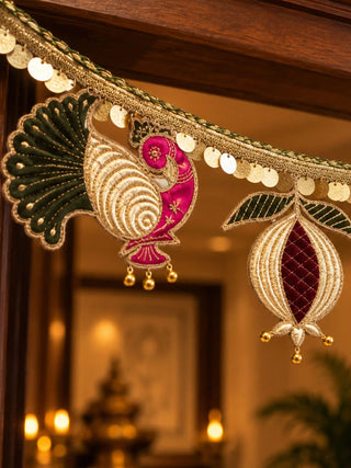 Close-up of the toran: a zari-worked peacock with a rani pink neck and green velvet tail beside a maroon velvet anar with gold bells.