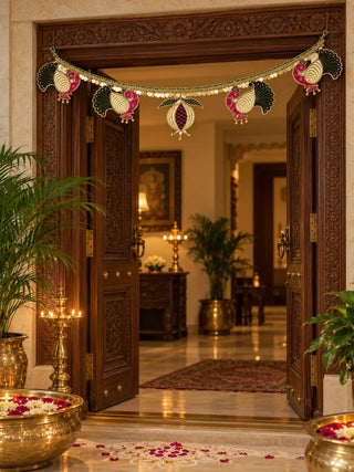 Mangal Mayura toran over an open carved door with brass diyas and a flower urli, pink and green velvet peacocks on a gold coin border.