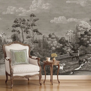 A black and white European vintage wallpaper featuring a manor estate scene with trees, horses, and a stone bridge, displayed behind a classic armchair and side table.
