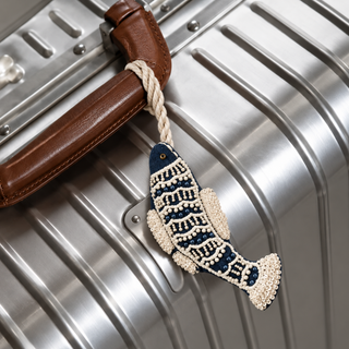 The Matsya Fish Charm in Navy & Ivory, tied to a suitcase handle by its braided cord, showing the beaded scale pattern and fin detail.