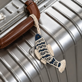 The Matsya Fish Charm in Navy & Ivory, tied to a suitcase handle by its braided cord, showing the beaded scale pattern and fin detail.
