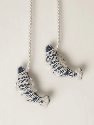 A studio view of the Matsya tieback featuring two indigo and silver hand-beaded fish charms on a cream cord.