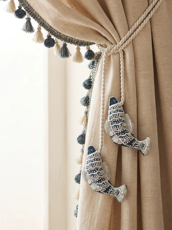 Hand-embroidered indigo fish charms with silver sequins holding back cream linen curtains with tassel trim.