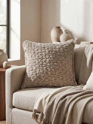 A lifestyle view of the Maya smocked velvet cushion styled on a light-colored armchair next to ceramic vases.