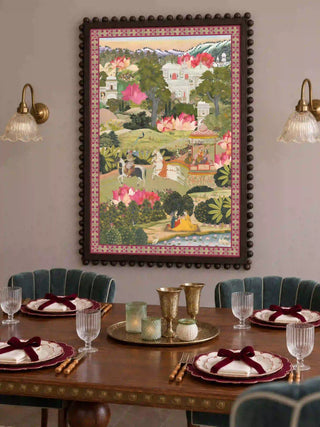 The Mewar wall art print in a dark beaded wood frame above a dining table set with burgundy bows, brass goblets and scalloped velvet chairs.