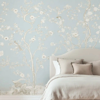 A light blue Chinoiserie wallpaper featuring delicate white floral blossoms and branches, styled behind a soft-toned bed with pillows.