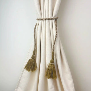 Mosswood Luxe Handcrafted Curtain Holdback secured around a cream curtain, showcasing the contrast of the olive tassels against the fabric.