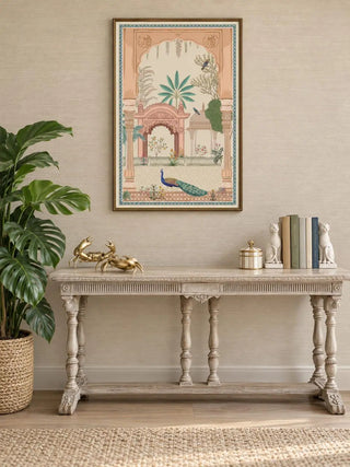 Mughal Garden portrait wall art in a thin gold-toned frame above a carved, pale distressed console table with brass crab ornaments, books between cat bookends and a monstera plant in a basket.