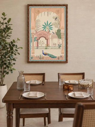 Mughal Garden portrait wall art in a wavy dark wood frame above a wooden dining table with cane-backed chairs, a glass carafe and amber candle holders.