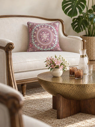 The Naqkashi embroidered cushion cover in mauve on a cream sofa with a carved wood frame, beside a hammered brass coffee table and pink flowers.