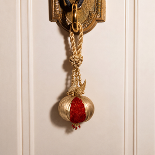 The Nar Pomegranate Hanging Charm in Gold & Red, hung from an ornate brass door handle by its braided cord and gold leaf detail.
