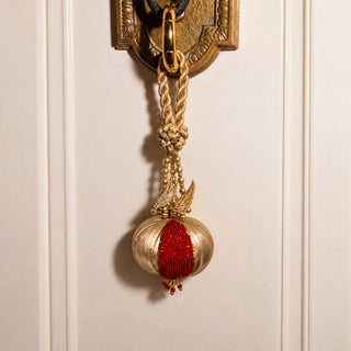 The Nar Pomegranate Hanging Charm in Gold & Red, hung from an ornate brass door handle by its braided cord and gold leaf detail.