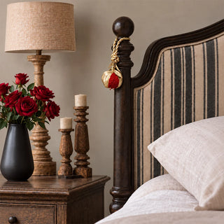 The Nar Pomegranate Hanging Gift Charm tied to a dark wooden bedpost beside a striped upholstered headboard and red roses.