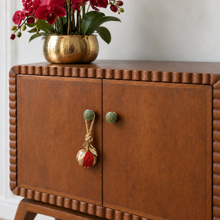 The Nar Pomegranate Hanging Gift Charm hung from a green knob on a warm brown wooden cabinet with a gold vase of red orchids.
