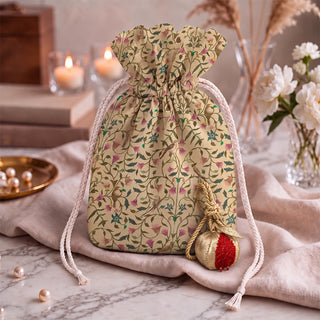 The Nar Pomegranate Hanging Gift Charm in gold and red resting beside a floral drawstring pouch with birds, on blush linen and marble.