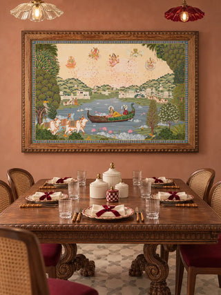 The Nauka-Vihar wall art in a carved wooden frame above a dark wood dining table with cane-back chairs on a terracotta wall.