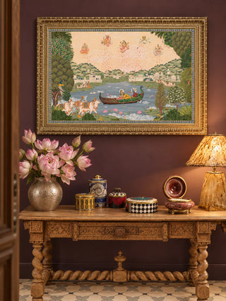 The Nauka-Vihar wall art in a gilt frame above a carved wooden console with pink flowers in a hammered vase, against a deep plum wall.