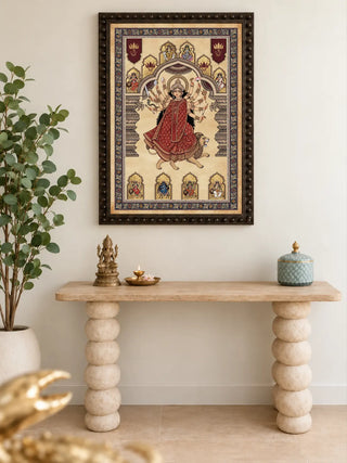 Framed Navadurga Virajitam wall art in a dark beaded frame above a travertine console with a brass figure, a brass lamp and a blue lidded jar