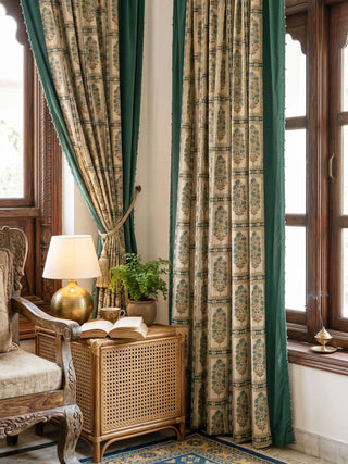 A pair of Nayab curtains framing a window in a traditional room with carved wood furniture and a brass lamp.