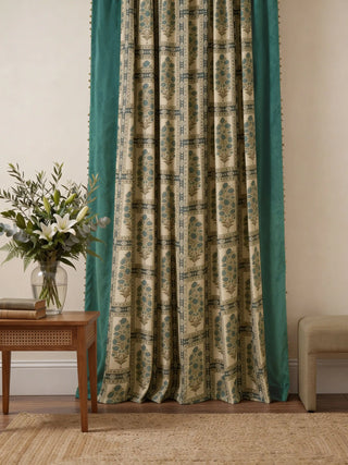 A full-length view of the Nayab curtain featuring teal botanical patterns, velvet borders, and gold coin fringe styled in a neutral room.