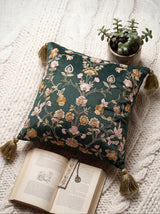 A luxury emerald green velvet cushion with hand-embroidered floral motifs and gold tassels styled with a book and plant.